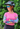 Woman wearing a colorful helmet, sunglasses, and arm sleeves with a pink shirt against a green leafy background.